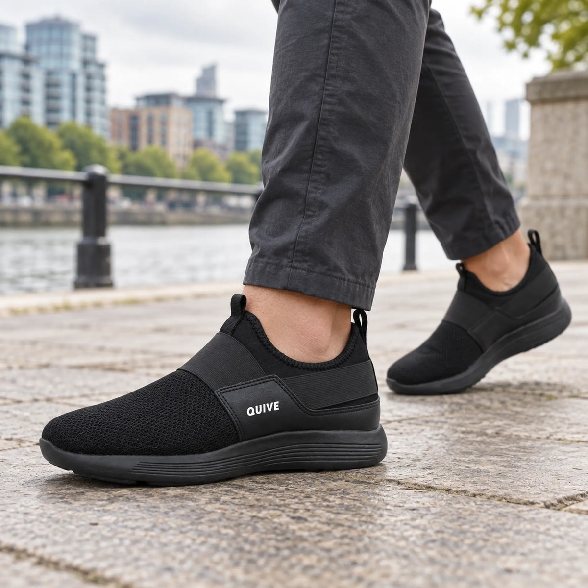 QUIVE black slip-on walking trainers worn on a British riverside promenade
